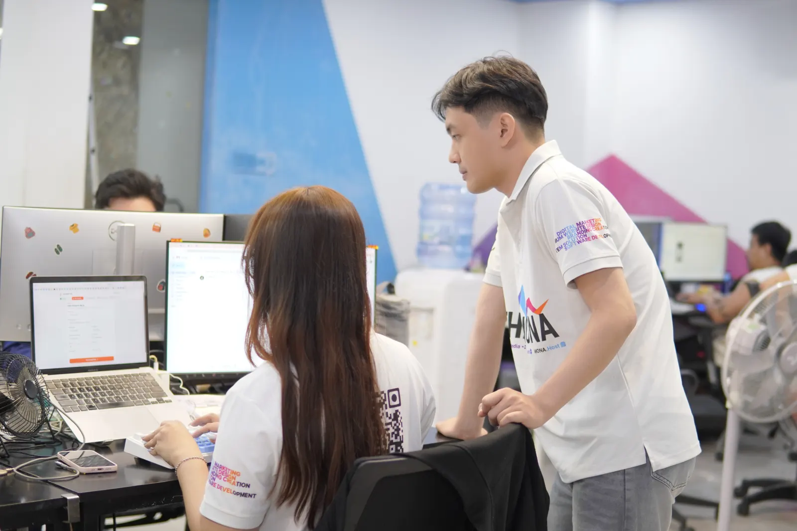 Anh đồng nghiệp đứng kèm chị đang làm trên laptop